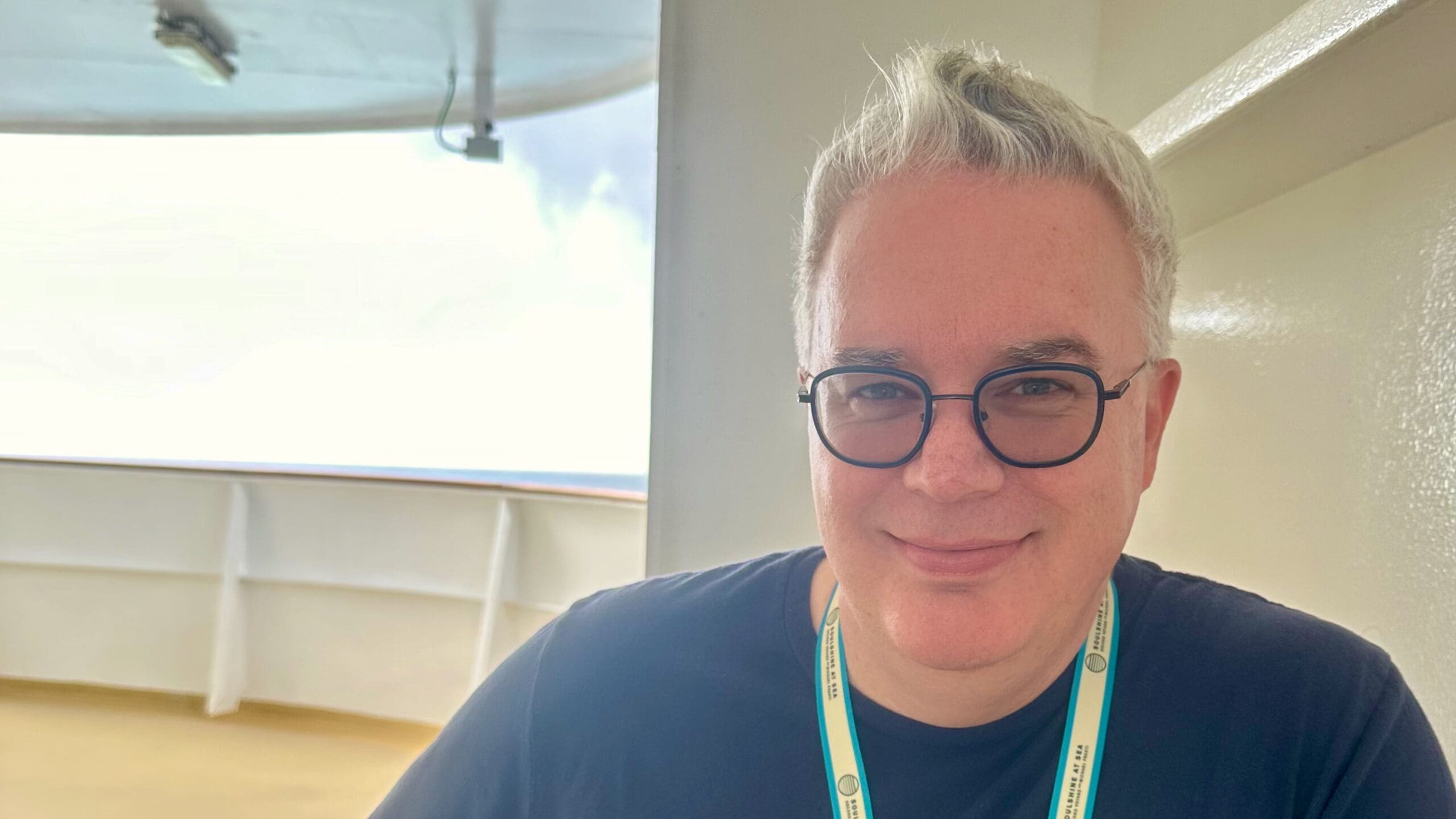 A white man with short, light-colored hair is sitting on a deck of a ship. The individual is wearing a black shirt and a lanyard around the neck. The background shows part of the ship's structure and an overcast sky.