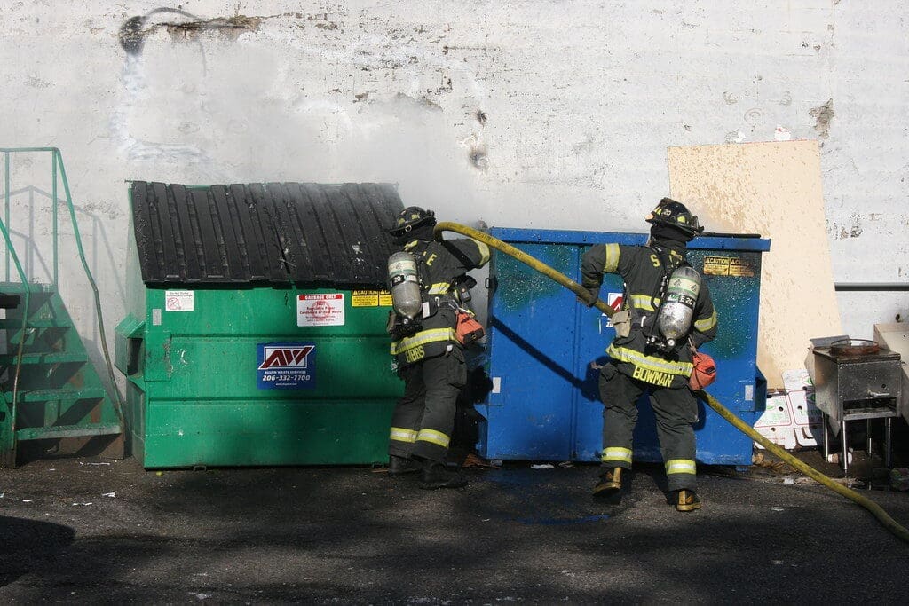 two firefighters use a long hose to extinguish a dumpster fire.